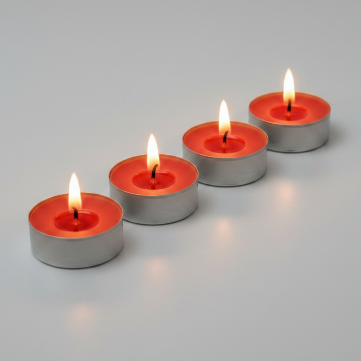 Four small tealight candles with lit flames arranged diagonally on a smooth light gray surface.