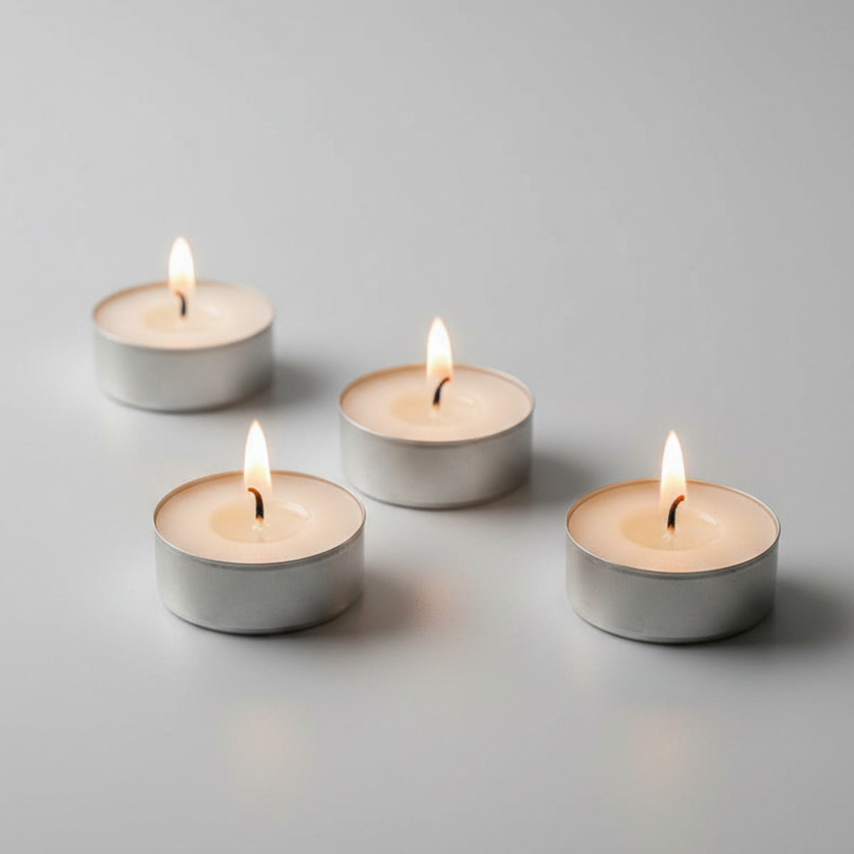 Four lit tealight candles in small silver metal cups arranged on a clean light gray surface against a soft gray background.