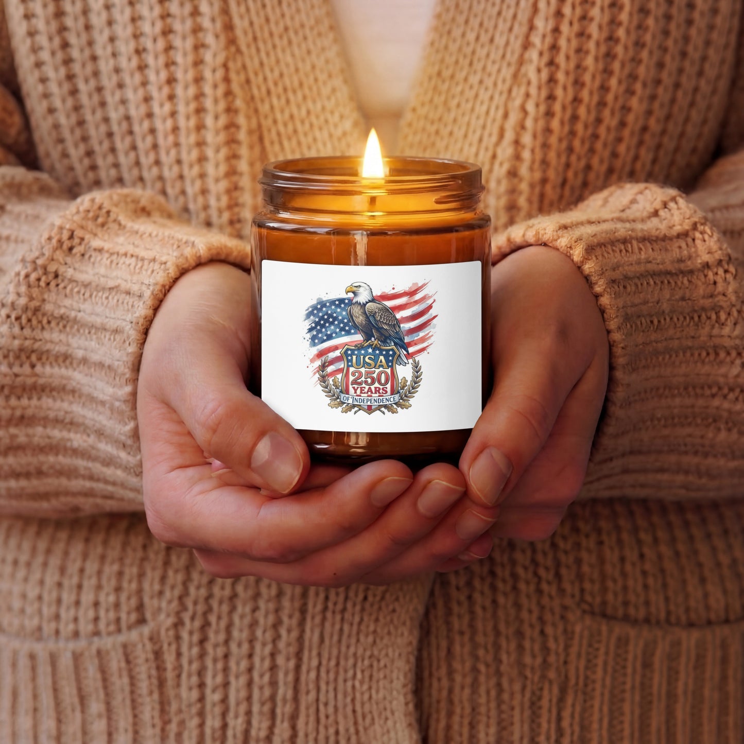 A lit amber glass jar candle held in a person's hands, wearing a beige knit sweater. The candle features a white label with an eagle and American flag graphic and the text 'USA 250 Years of Independence'.