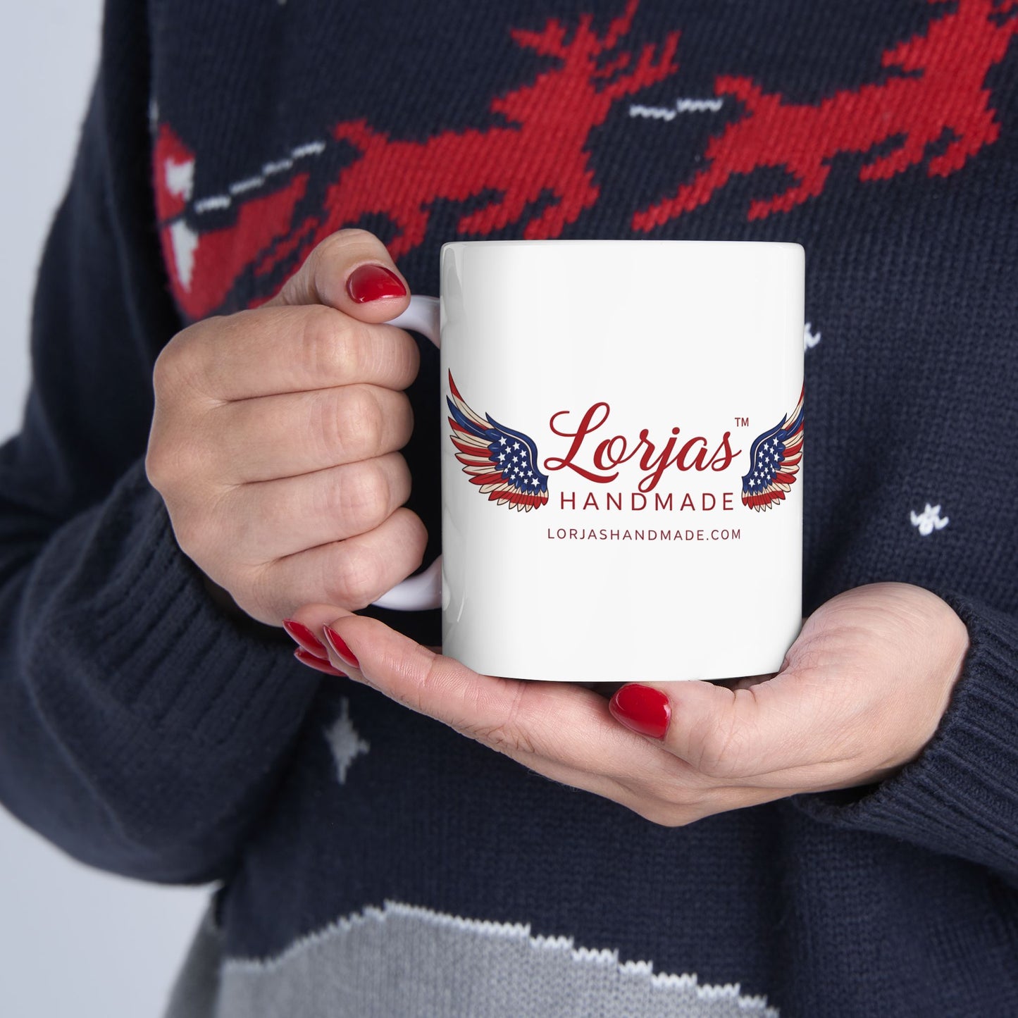 Person holding a white mug with 'Lorjas Handmade' logo, wearing a dark sweater with red deer design.
