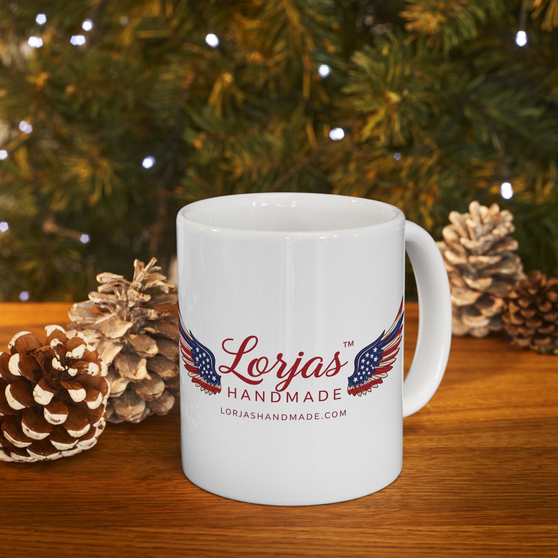 White mug with 'Lorjas Handmade' logo in front of a Christmas tree and pinecones.