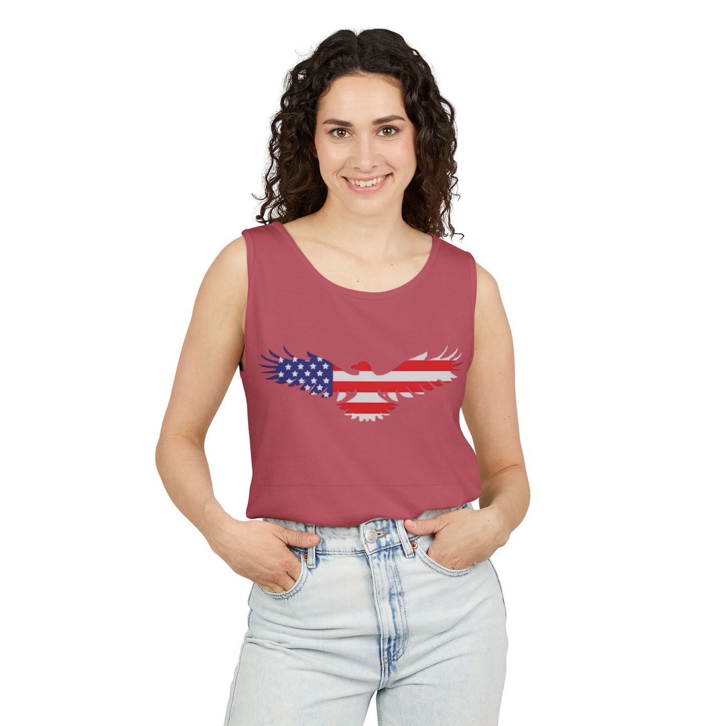 A woman modeling a sleeveless scoop-neck tank top in dusty rose pink, featuring a distressed eagle graphic filled with the American flag pattern (stars and stripes) on the chest. She is wearing light-wash blue jeans against a solid white background.