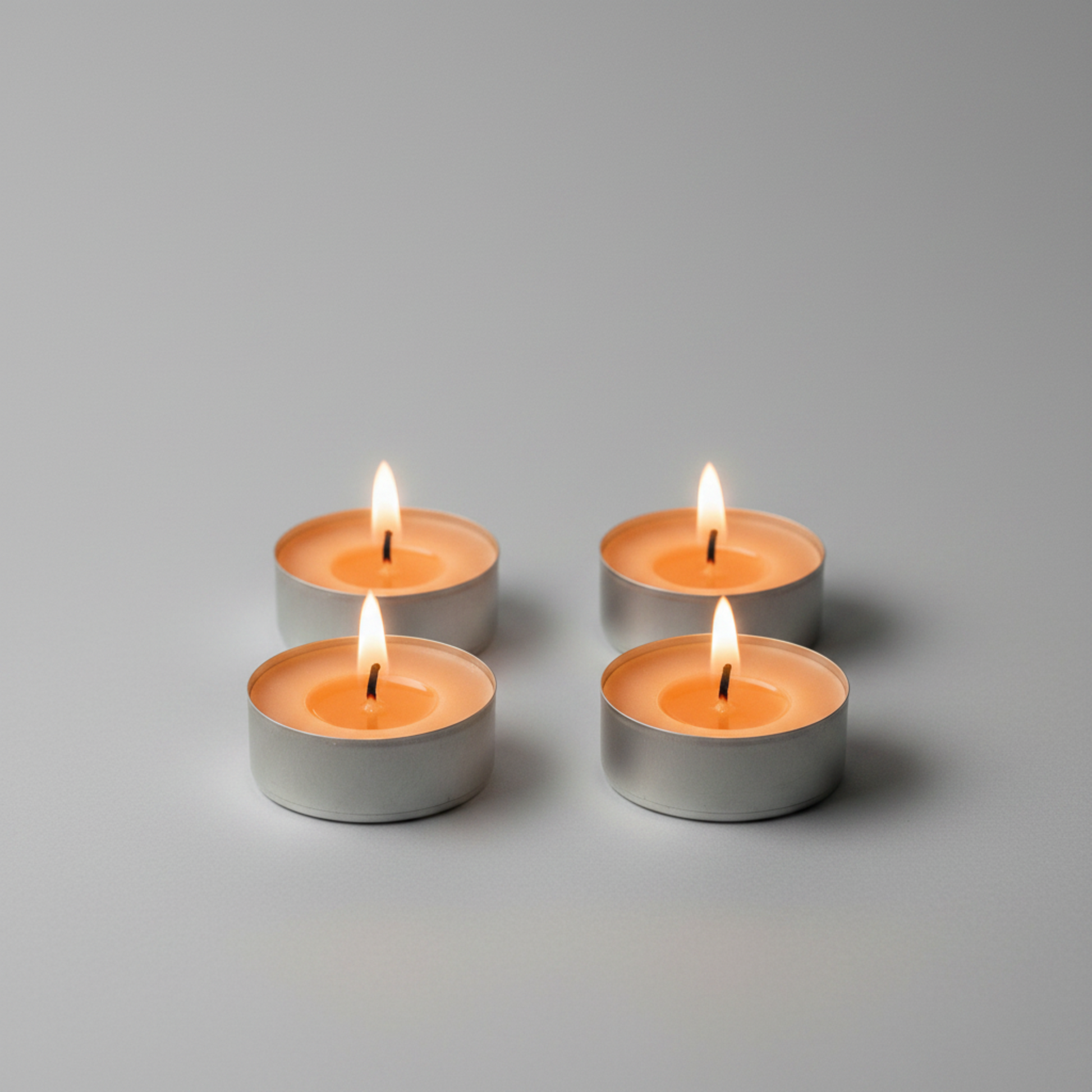 Four lit tealight candles in small silver metal holders arranged in a 2x2 grid on a smooth light gray surface with a soft gray background.