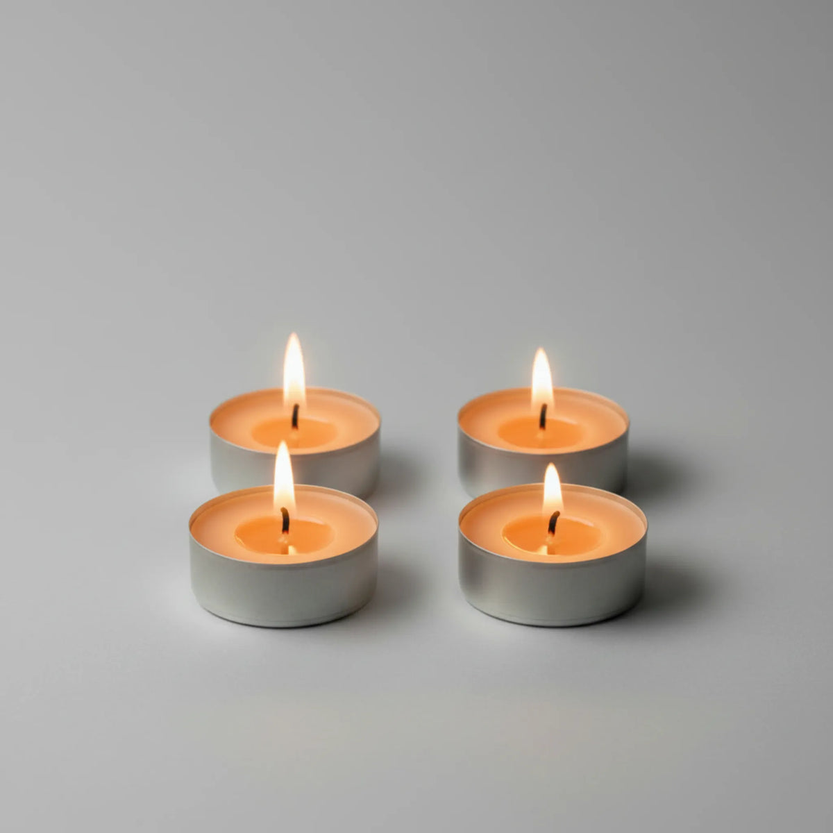 Four lit tealight candles in small silver metal holders arranged in a 2x2 grid on a smooth light gray surface with a soft gray background.