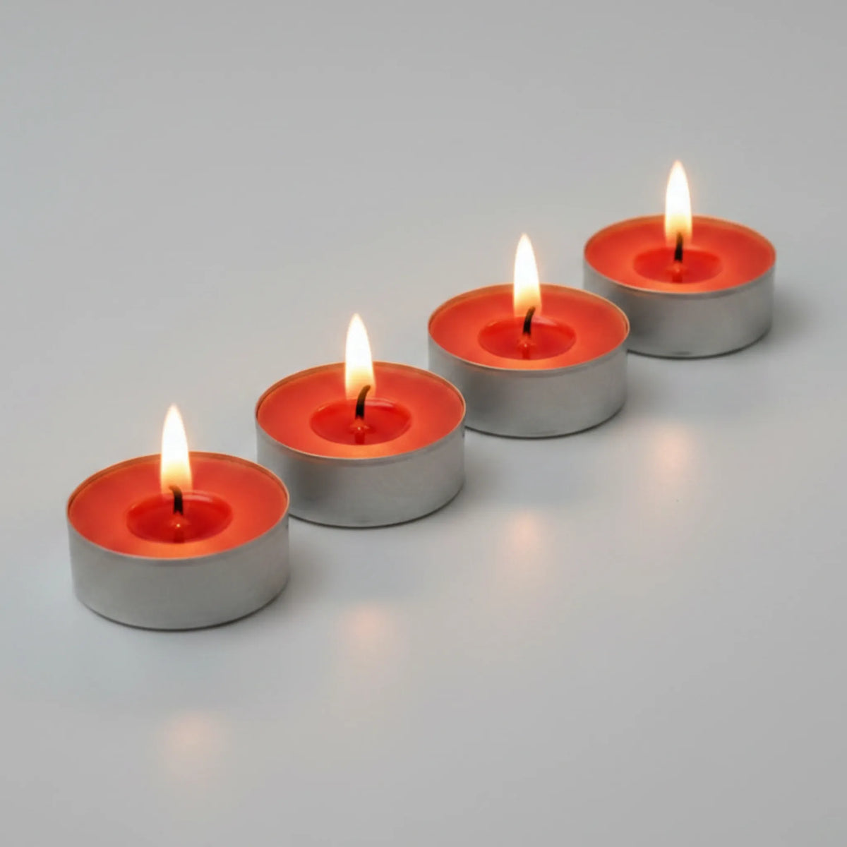 Four small tealight candles with lit flames arranged diagonally on a smooth light gray surface.