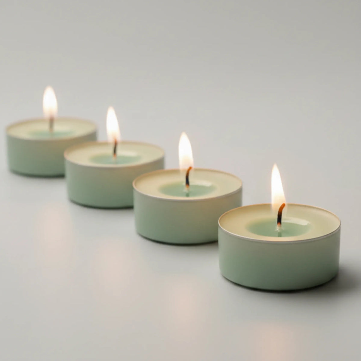 Four small tealight candles in pale mint green holders, arranged in a diagonal line with their flames lit, on a smooth light gray surface.