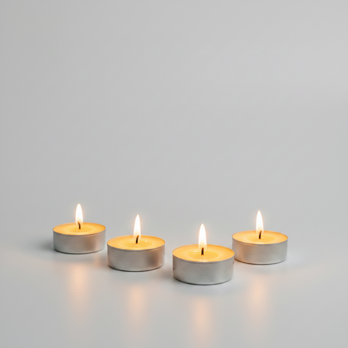 Four lit silver tea light candles arranged in a row on a light gray surface against a matching gray background.