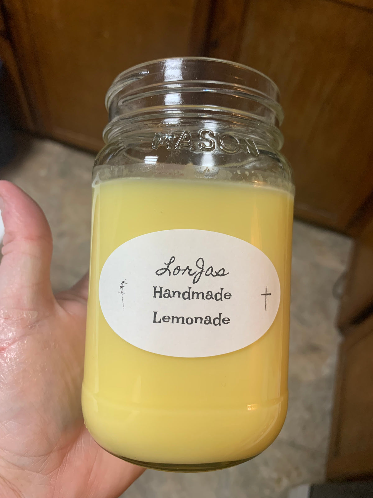 LorJas Handmade Lemonade scented soy wax candle held in a hand with a blurred tiled background and kitchen shelving.