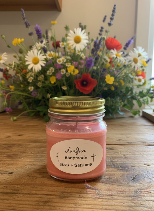 Pink yuzu & satsuma candle in a jar with a gold lid on a wooden surface, surrounded by a bouquet of flowers.