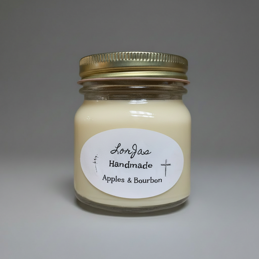 A handmade candle in a clear glass jar with a gold metal screw-on lid and a white oval label reading 'Lor’Jas Handmade Apples & Bourbon.' The candle wax is a warm cream color, photographed against a neutral gray studio background.