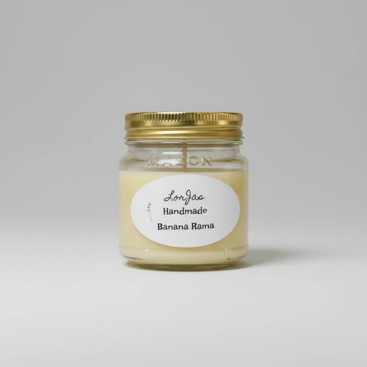 A handcrafted candle in a clear glass mason jar with a gold metal lid and a white oval label reading 'Lor'Jas Handmade Banana Rama', centered on a light gray background.