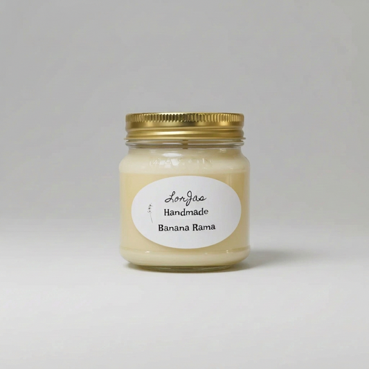 A handcrafted candle in a clear glass mason jar with a gold metal lid and a white oval label reading 'Lor'Jas Handmade Banana Rama', centered on a light gray background.
