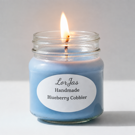 A pleasantly lit Lor’Jas Handmade ‘Blueberry Cobbler’ lightly scented soy wax candle in a glass jar with light blue wax and a gold metal lid, standing on a white surface against a light gray background.