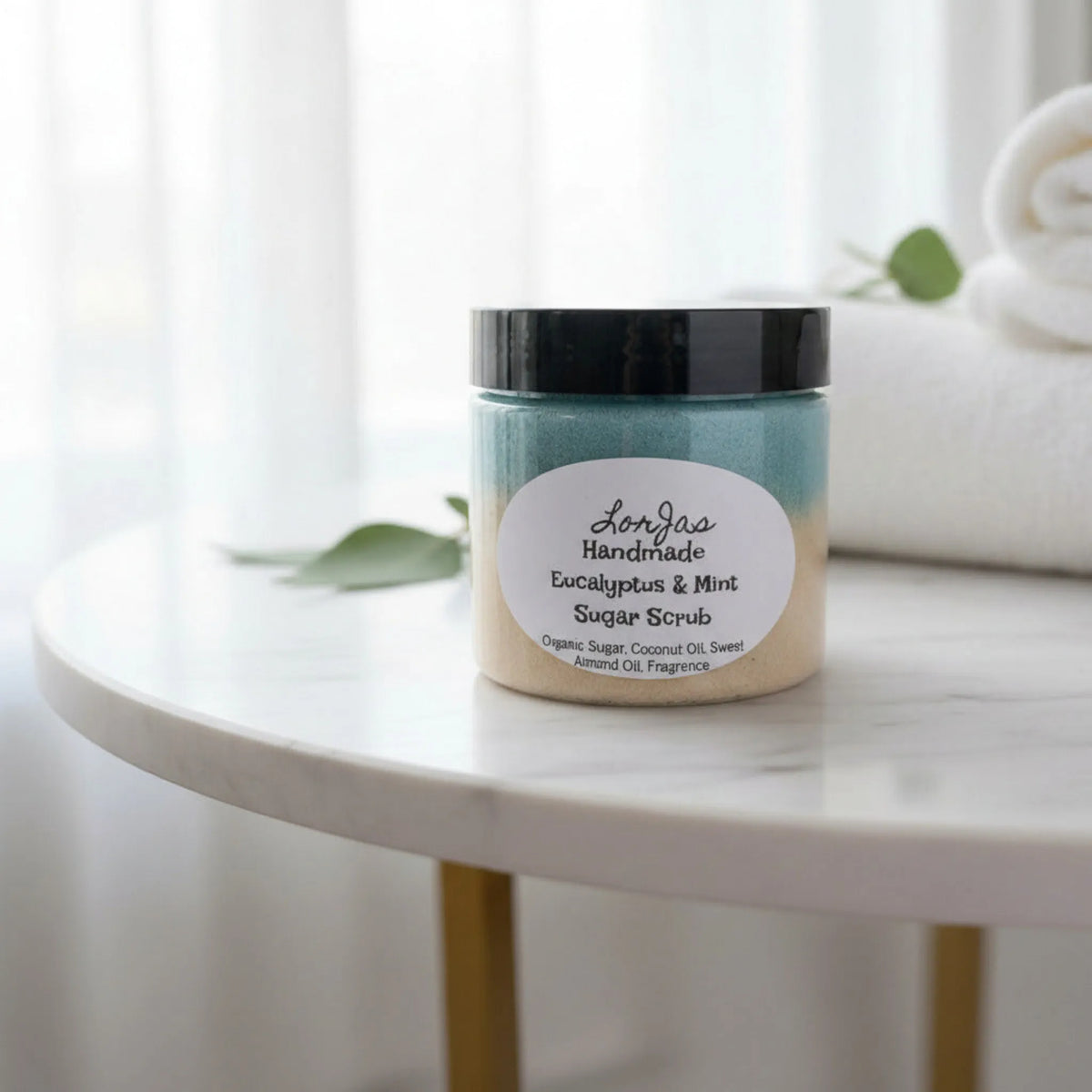 LorJas Handmade lightly scented Eucalyptus & Mint sugar scrub on a marble table with rolled towels by a draped window letting natural light flow through in a spa setting. 