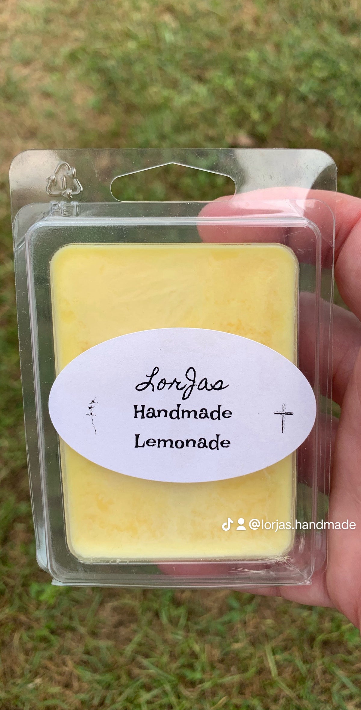 Yellow wax melt in packaging with 'Lorjas Handmade Lemonade' label held by a hand against a grassy background.