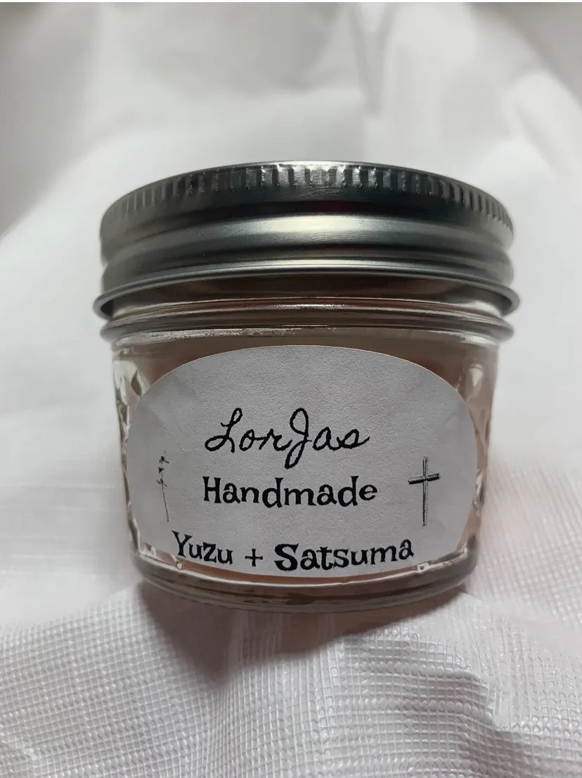 A small clear glass jar with a silver metal screw lid, containing a scented soy candle, with a white oval label that reads 'Lorjas Handmade Yuzu + Satsuma'. The jar is placed on a white textured fabric background.
