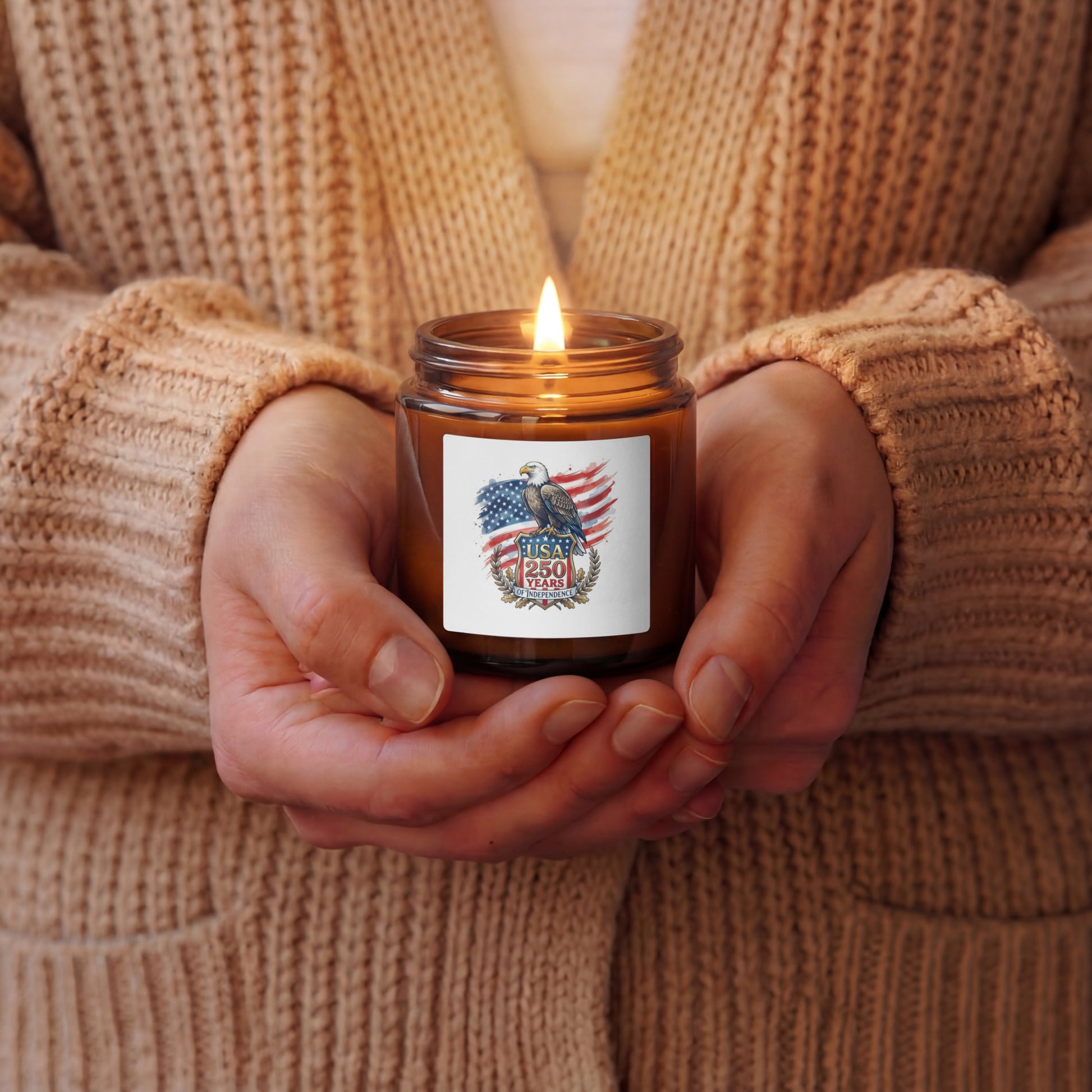 A person in a beige knit cardigan holds a lit amber glass jar candle with a white label featuring an eagle and American flag design. The candle is shown in a cozy, warm setting.