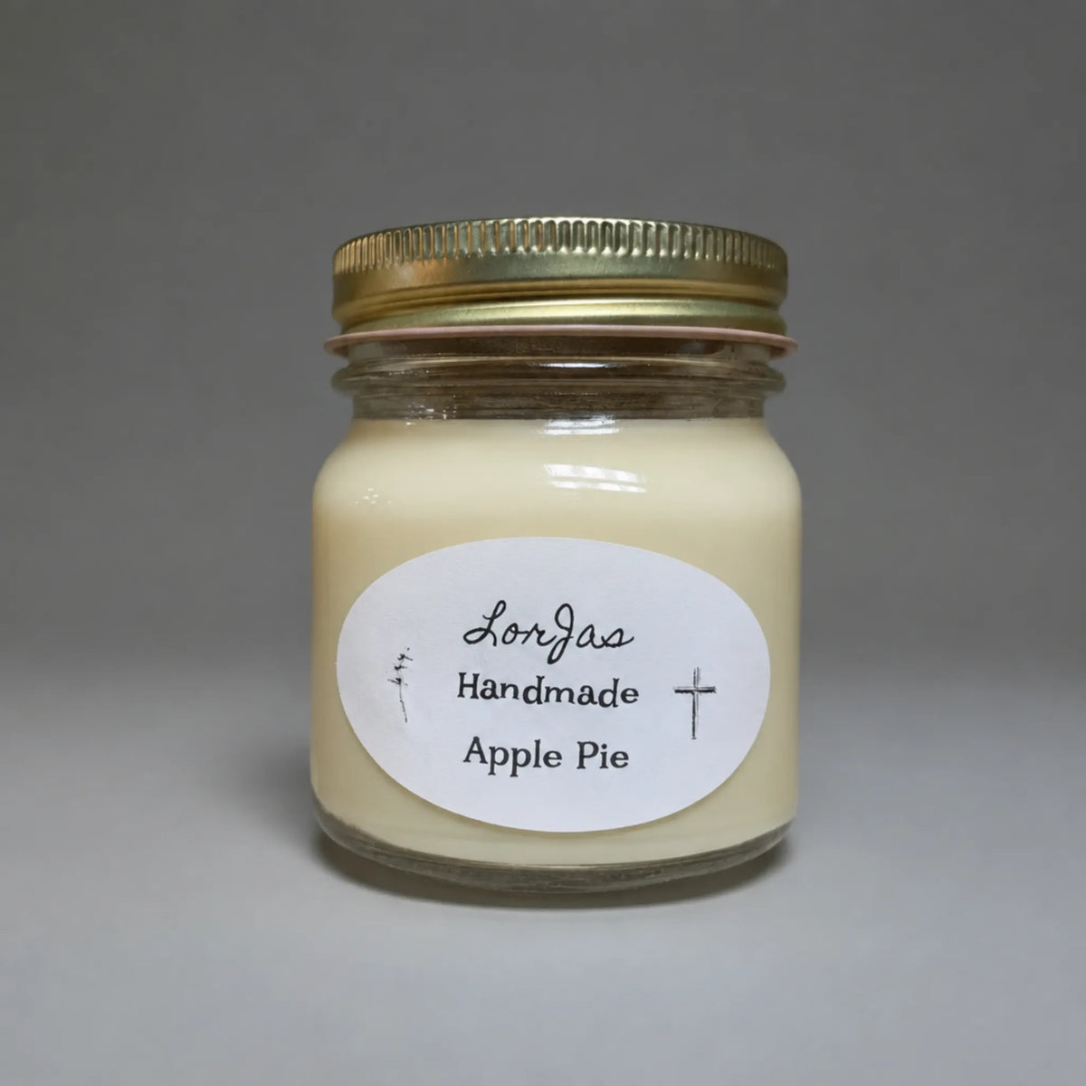 A glass jar candle with creamy off-white wax, a gold metal screw-top lid, and a white oval label reading 'LorJas Handmade Apple Pie' on a neutral gray background.