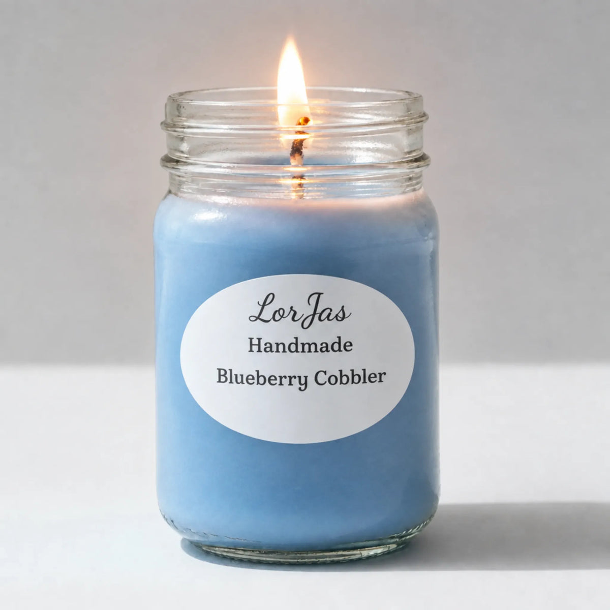 A lit blue candle in a clear glass mason jar with a white oval label reading 'LorJas Handmade Blueberry Cobbler' on a light gray background.