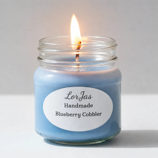 A lit blue candle in a clear glass jar with a white oval label reading 'LorJas Handmade Blueberry Cobbler' on a light gray background.