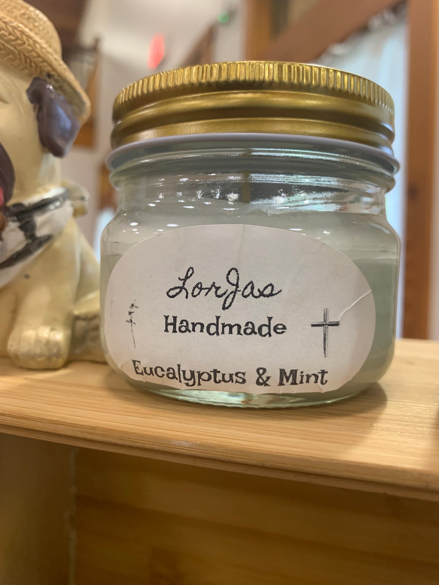 LorJas Handmade 'Eucalyptus & Mint' scented organic soy candle in glass jar with a gold lid and a branded label on top of a wooden display at market beside furry friend caricature.