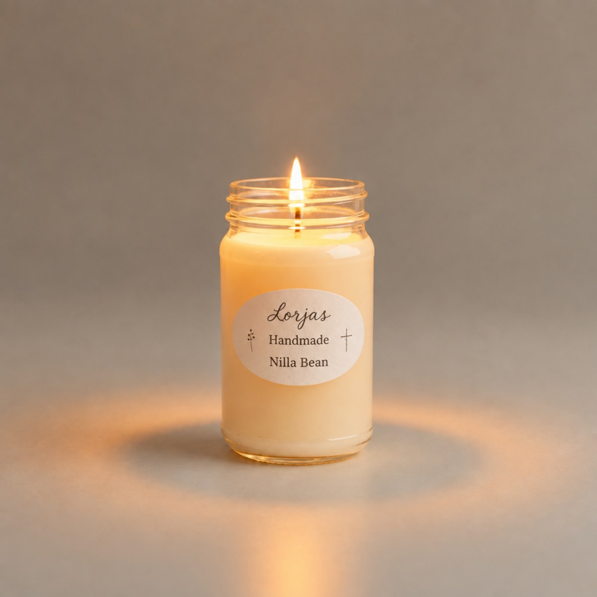 A lit jar candle with creamy off-white wax in a clear glass jar, featuring a white oval label that reads 'Lor'Jas Handmade Nilla Bean' on a neutral gray background.