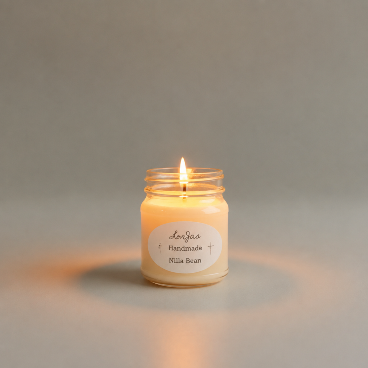 A lit candle in a clear glass jar with a white label reading 'Lor'Jas Handmade Nilla Bean,' centered on a soft warm gray background.