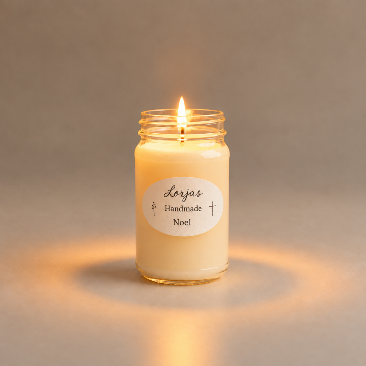 A lit candle in a clear glass jar labeled 'Lor’Jas Handmade Noel,' centered on a soft warm gray background.