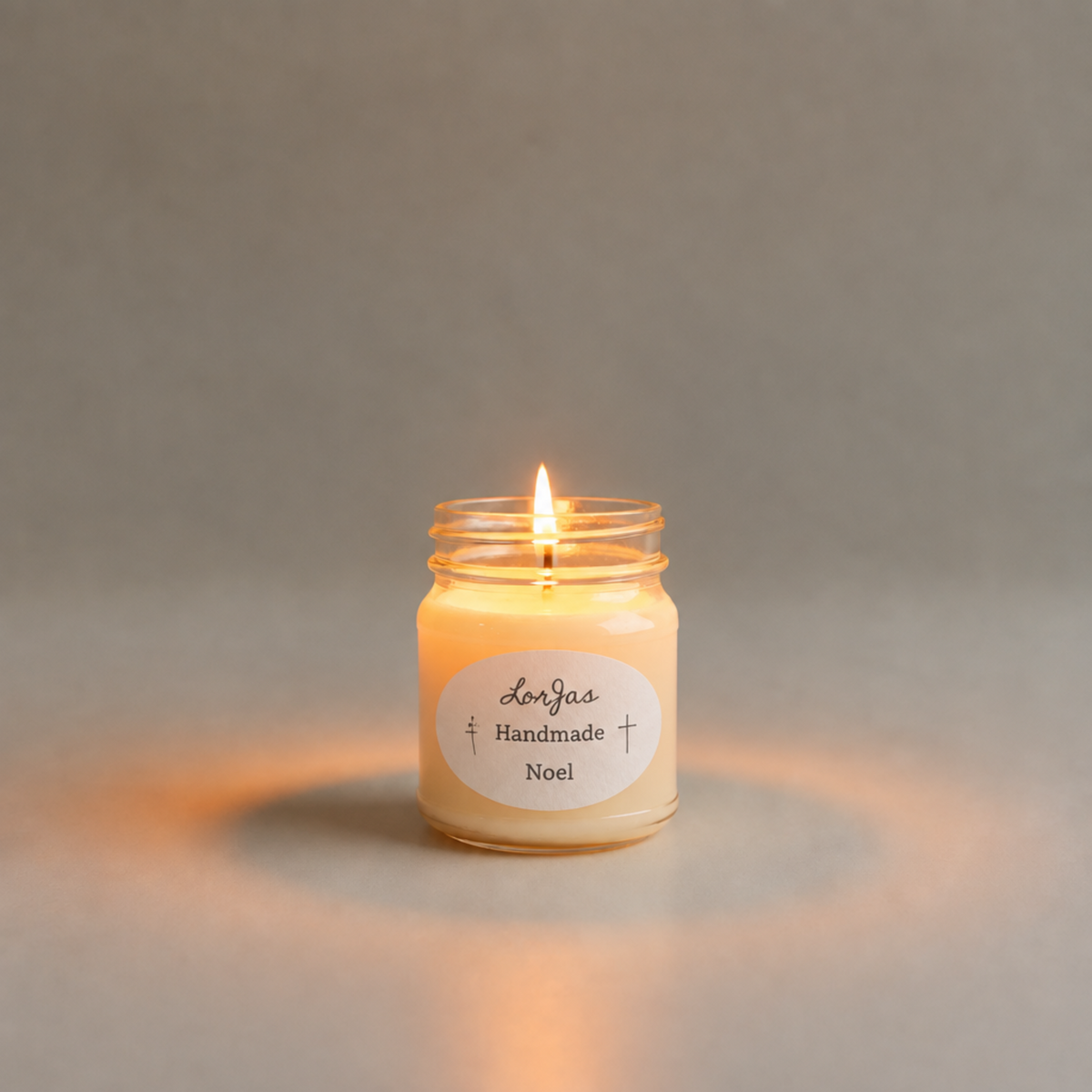 A lit candle in a clear glass jar labeled 'Lor’Jas Handmade Noel,' centered on a soft warm gray background.