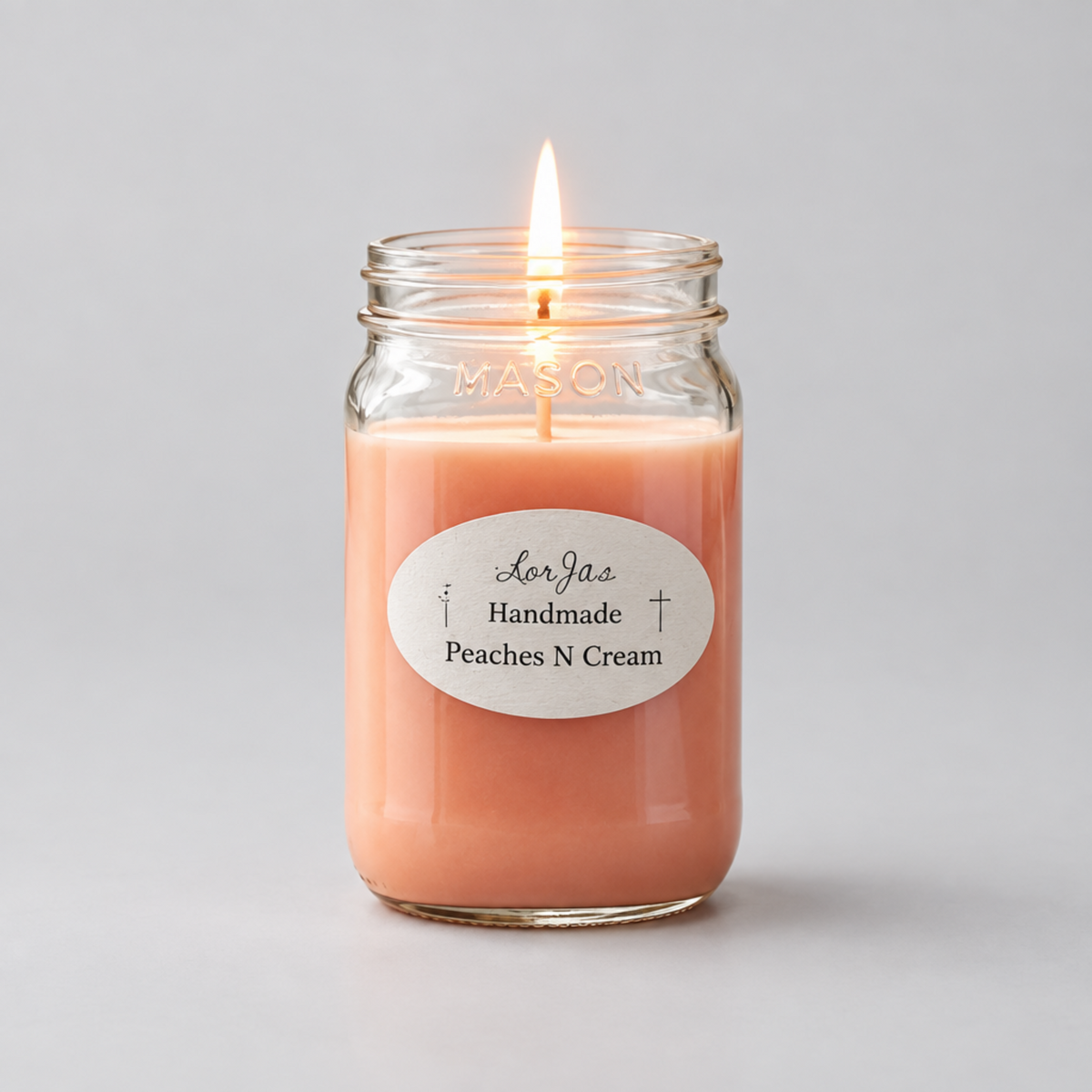 A lit mason jar candle with peach-colored wax, featuring a white oval label that reads 'Lor'Jas Handmade Peaches N Cream' on a neutral light gray background.