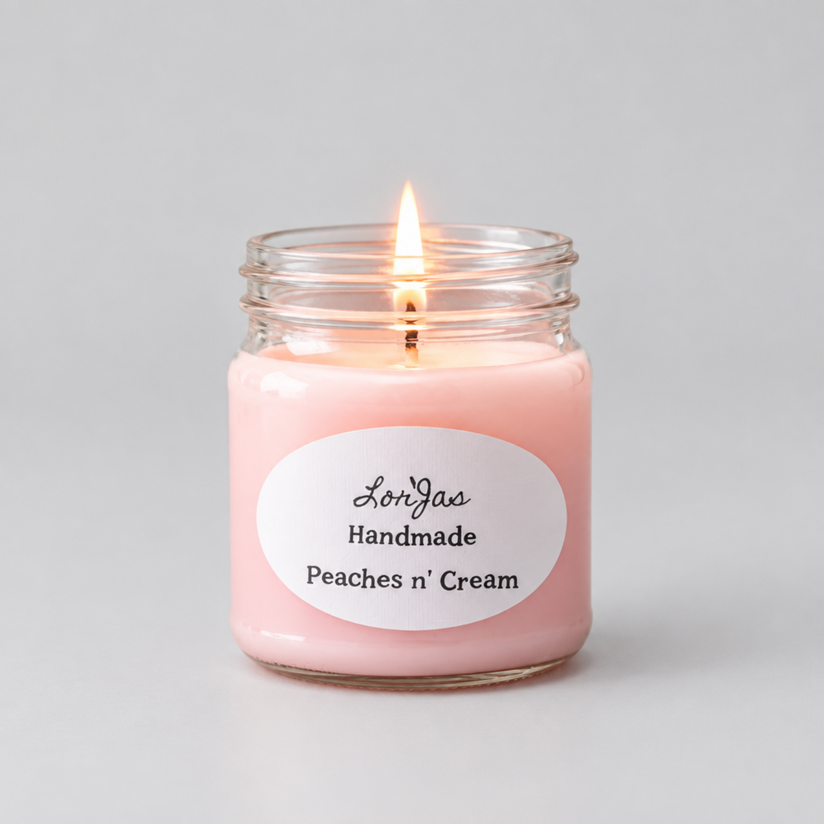 A lit pink jar candle in a clear glass mason jar with a white oval label reading 'Lor'Jas Handmade Peaches n' Cream' on a light gray background.
