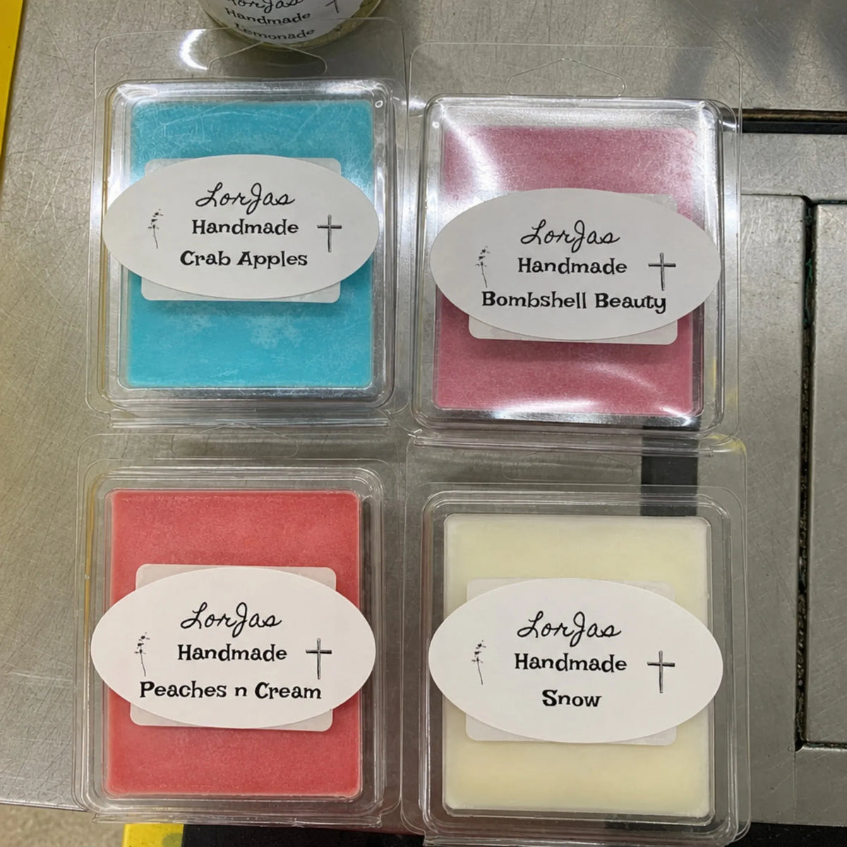 Four square wax melts in clear plastic clamshell packaging arranged in a 2x2 grid on a stainless steel surface. The labels read LorJas Handmade and list Crab Apples, Bombshell Beauty, Peaches n Cream, and Snow in different pastel and bright colors: blue, pink, coral-red, and off-white.
