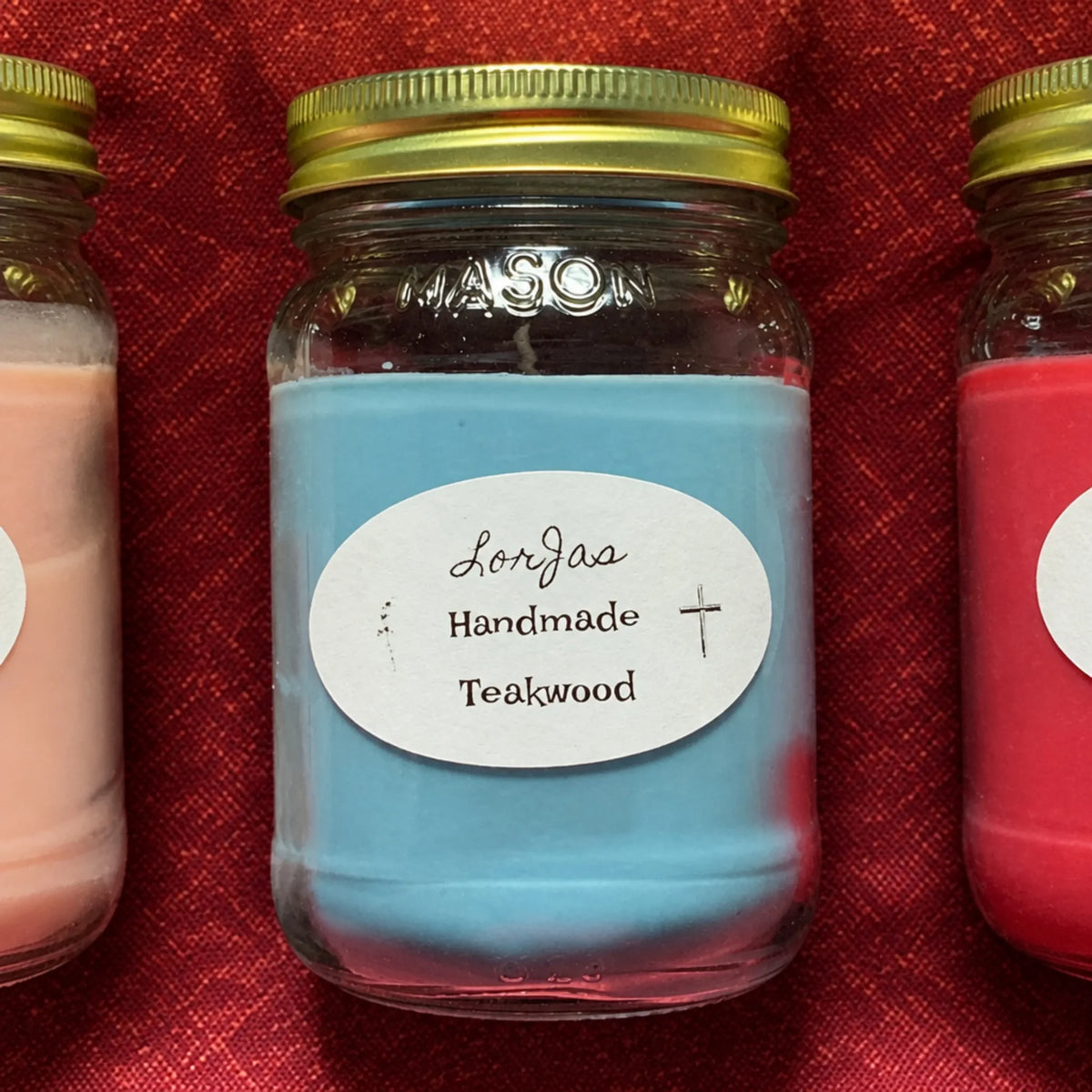 A LorJas Handmade teakwood candle in a clear glass mason jar with a gold metal lid, featuring a light blue wax body and a white oval label that reads 'LorJas Handmade Teakwood'. The jar is displayed on a textured red fabric next to two other jars partially visible on the left and right.