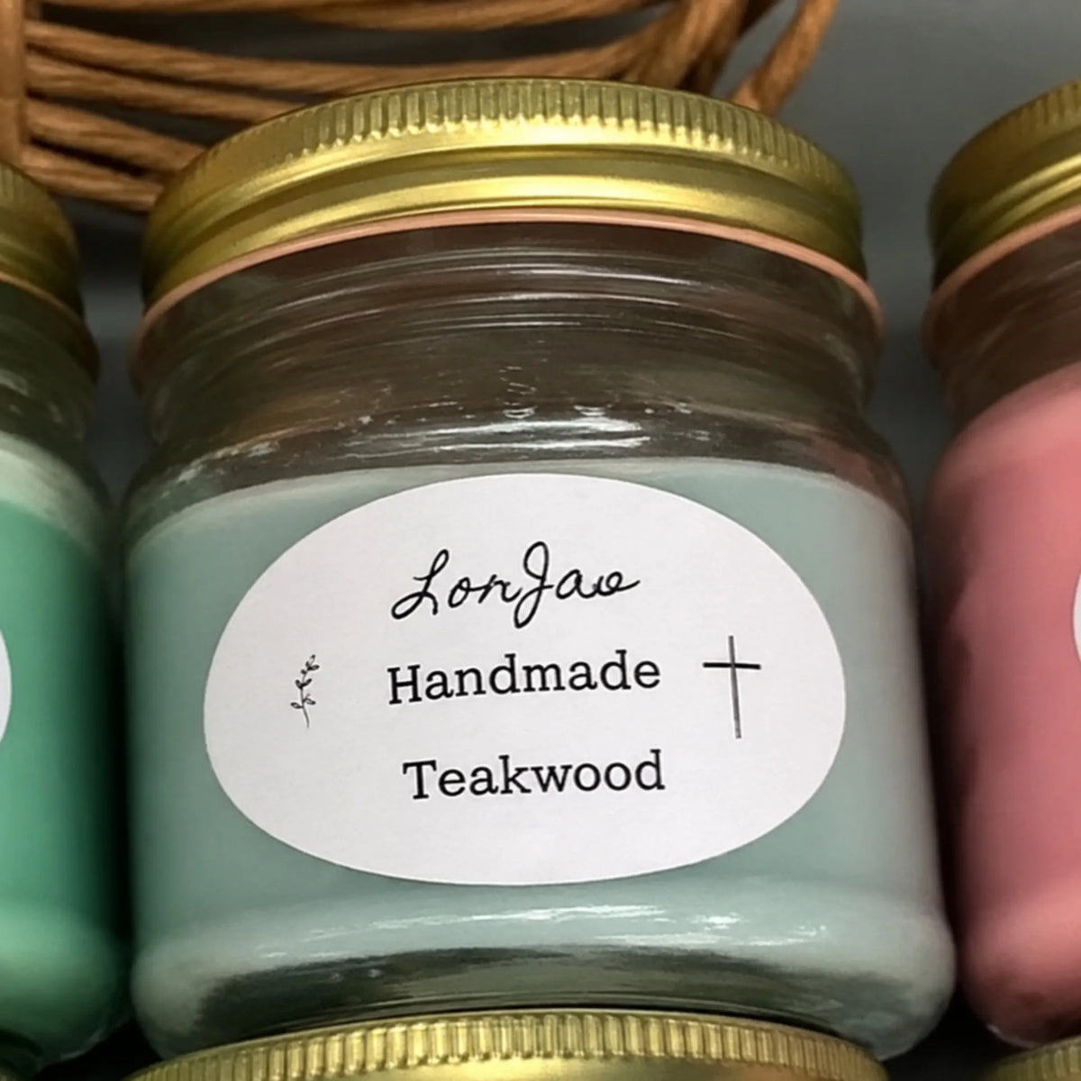 A close-up of a LorJas Handmade teakwood scented candle in a clear glass jar with a gold metal lid and a white oval label reading 'LorJas Handmade Teakwood'.