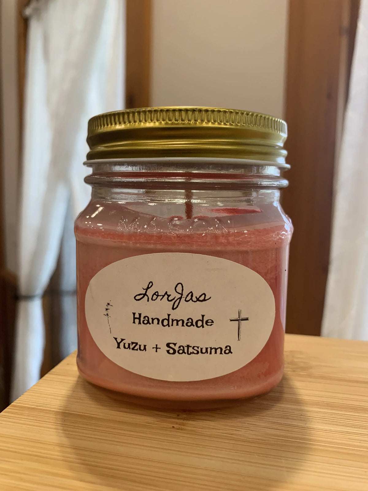A handmade jar candle with pink wax in a clear glass jar, featuring a gold metal lid and a white oval label that reads 'LorJas Handmade Yuzu + Satsuma'. The candle is placed on a light wooden table with a softly blurred indoor background.